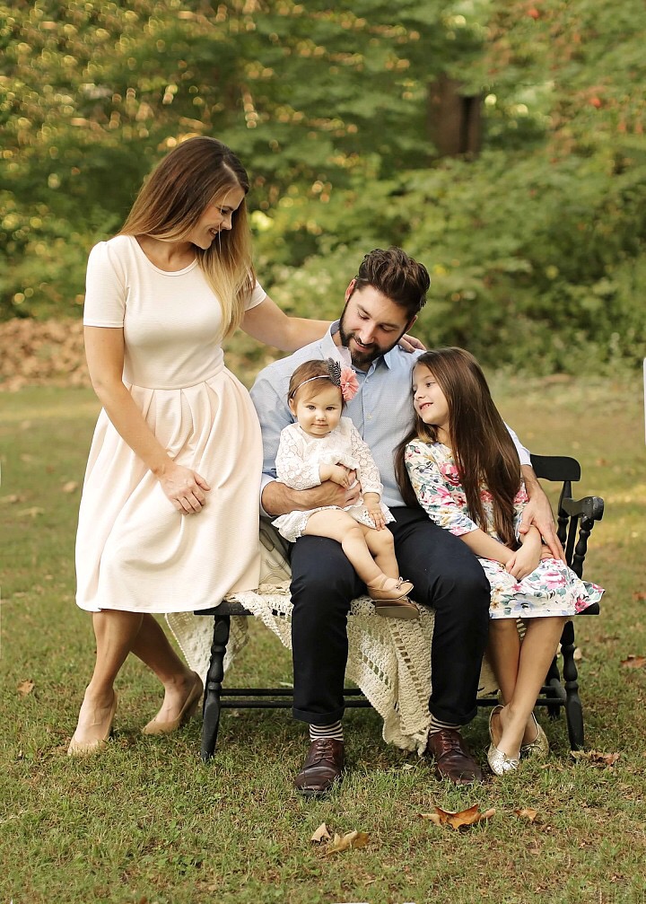 Katie Dunlavy, Lularoe boutique owner, of The Pearl Pages wearing a white dress and posing outside with her husband and two daughters sitting on a black bench.
